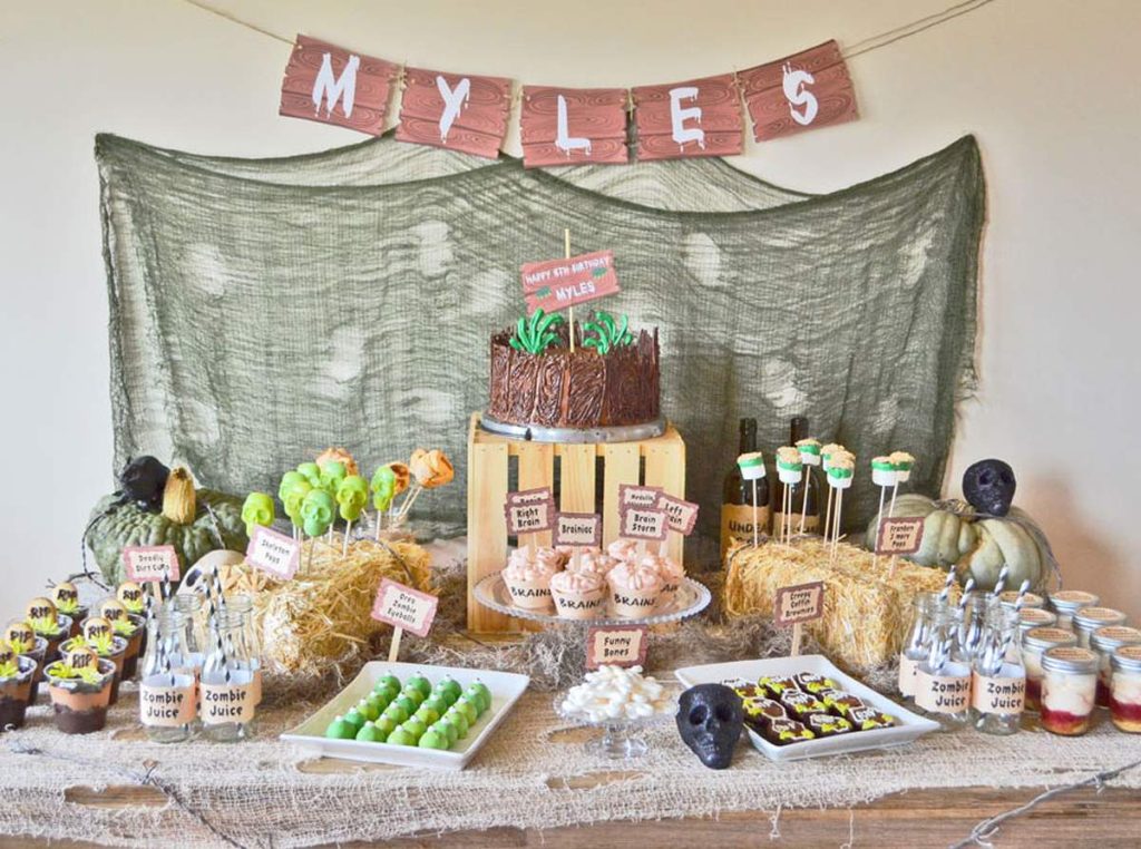 a festive and elaborately decorated "Zombie Apocalypse" themed birthday party dessert table for "Myles," featuring a variety of themed treats and decorations. The table is draped with a distressed, green-hued fabric resembling netting, and a banner spelling "MYLES" hangs above.